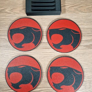 May include: Four round coasters with a red background and a black panther head design. A black rectangular holder with horizontal slots is above the coasters. The coasters are on a light brown wooden surface.