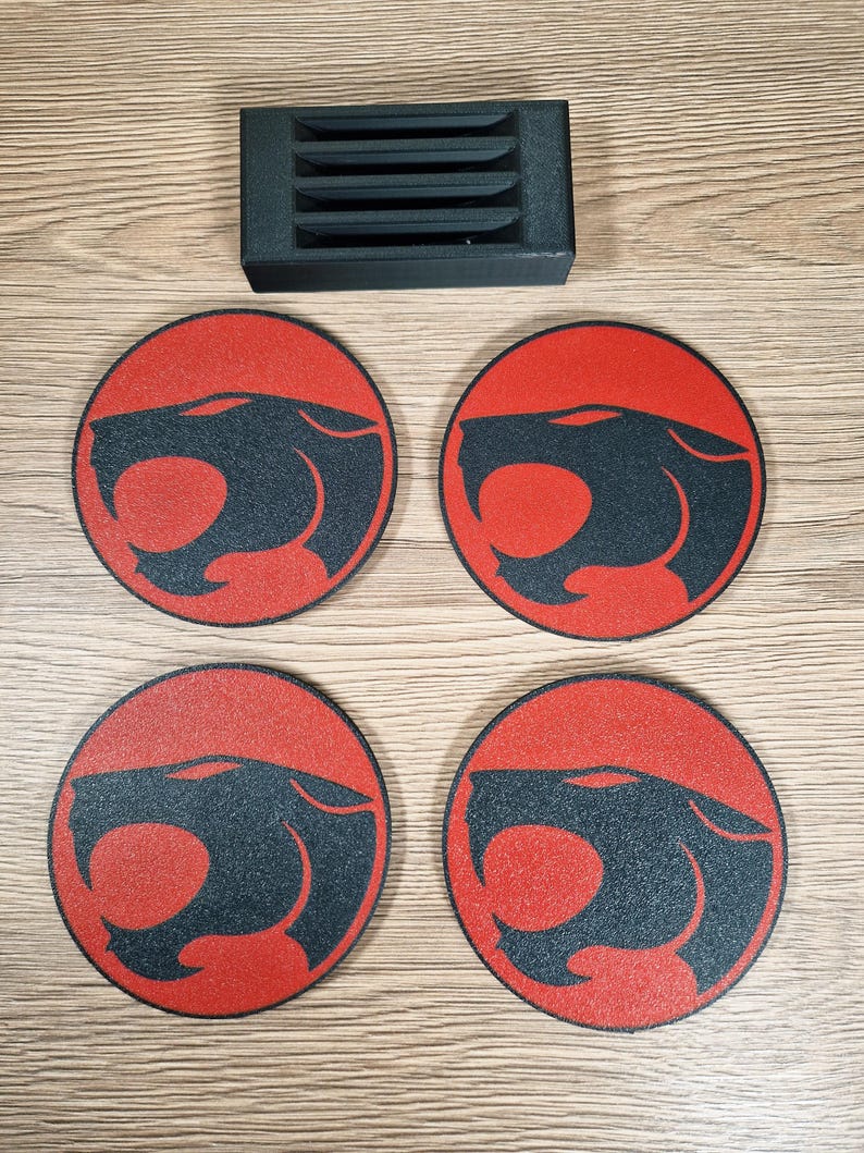 May include: Four round coasters with a red background and a black panther head design. A black rectangular holder with horizontal slots is above the coasters. The coasters are on a light brown wooden surface.