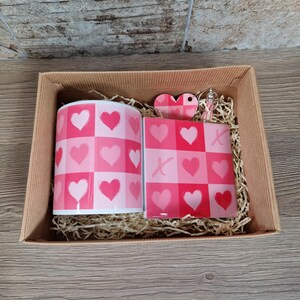 May include: A gift set in a cardboard box, featuring a mug and a coaster with a pink heart pattern. A heart-shaped keychain is also included. The mug and coaster have a checkerboard design with pink hearts.