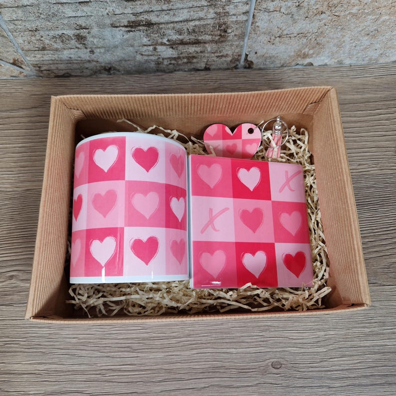 May include: A gift set in a cardboard box, featuring a mug and a coaster with a pink heart pattern. A heart-shaped keychain is also included. The mug and coaster have a checkerboard design with pink hearts.