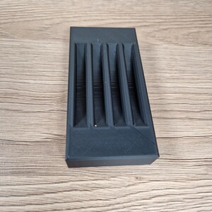 K&ouml;nnte beinhalten: Ein rechteckiger schwarzer Kunststoff-Organizer mit vier parallelen vertikalen Schlitzen. Das Objekt hat eine matte Oberfl&auml;che mit sichtbaren 3D-Druckschichten, die ein strukturiertes und funktionales Design zur Aufbewahrung kleiner Gegenst&auml;nde bieten.