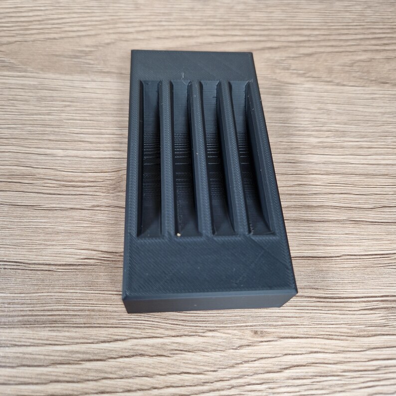 K&ouml;nnte beinhalten: Ein rechteckiger schwarzer Kunststoff-Organizer mit vier parallelen vertikalen Schlitzen. Das Objekt hat eine matte Oberfl&auml;che mit sichtbaren 3D-Druckschichten, die ein strukturiertes und funktionales Design zur Aufbewahrung kleiner Gegenst&auml;nde bieten.