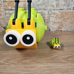 Puede incluir: Un caracol de juguete grande, amarillo y verde, con antenas negras y grandes ojos con borde blanco. Tambi&eacute;n hay una versi&oacute;n m&aacute;s peque&ntilde;a del caracol. Los juguetes son de pl&aacute;stico y est&aacute;n sobre una superficie de madera.