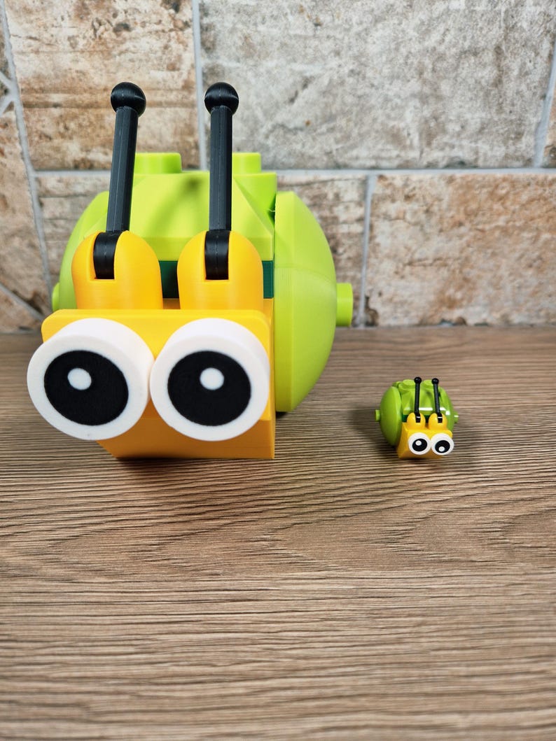 Puede incluir: Un caracol de juguete grande, amarillo y verde, con antenas negras y grandes ojos con borde blanco. Tambi&eacute;n hay una versi&oacute;n m&aacute;s peque&ntilde;a del caracol. Los juguetes son de pl&aacute;stico y est&aacute;n sobre una superficie de madera.