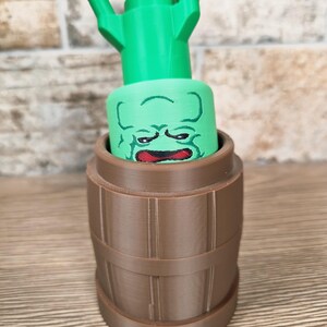 May include: A green watering can with a cartoon face design, set inside a brown barrel-shaped container. The watering can has a long spout and a handle. The face is green with red lips and a grumpy expression. The barrel has vertical lines.