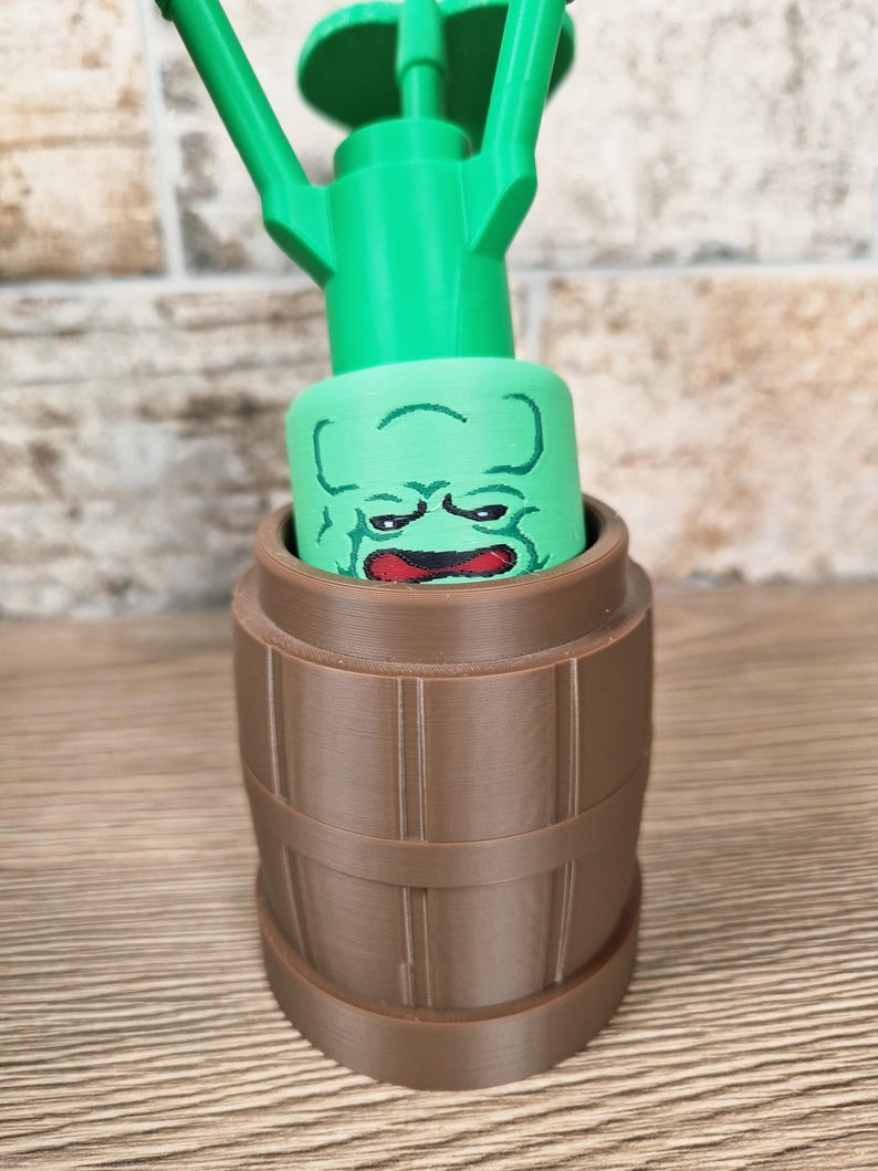 May include: A green watering can with a cartoon face design, set inside a brown barrel-shaped container. The watering can has a long spout and a handle. The face is green with red lips and a grumpy expression. The barrel has vertical lines.