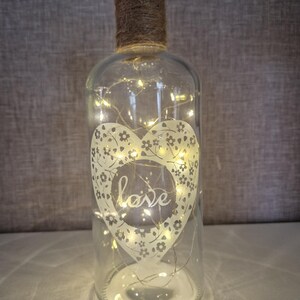 May include: A clear glass bottle with a heart design etched on the front. The heart has a floral pattern and the word "love" inside. The bottle is filled with white fairy lights.