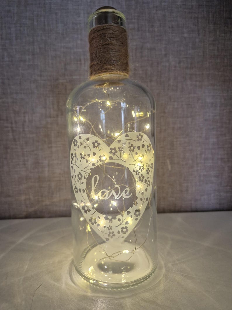May include: A clear glass bottle with a heart design etched on the front. The heart has a floral pattern and the word "love" inside. The bottle is filled with white fairy lights.