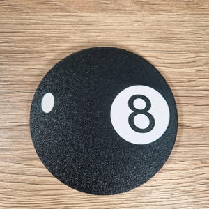 May include: A round, black coaster with a textured surface. The coaster features a white circle with the number 8 in the center, and a smaller white circle on the left side. The coaster is designed to resemble the 8-ball from a game of pool.
