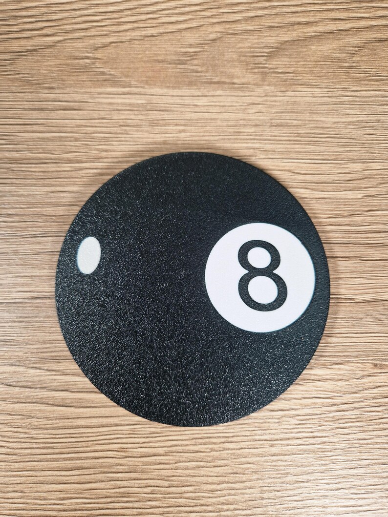 May include: A round, black coaster with a textured surface. The coaster features a white circle with the number 8 in the center, and a smaller white circle on the left side. The coaster is designed to resemble the 8-ball from a game of pool.