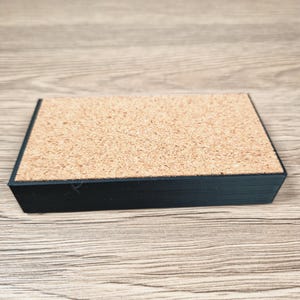 Puede incluir: Un posavasos rectangular de corcho con borde negro. El posavasos tiene una superficie de corcho natural y un borde negro ligeramente elevado. Est&aacute; dise&ntilde;ado para proteger las superficies del calor y la humedad.