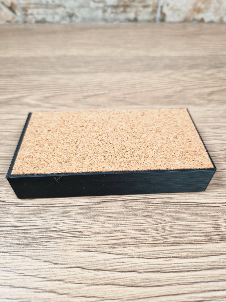Puede incluir: Un posavasos rectangular de corcho con borde negro. El posavasos tiene una superficie de corcho natural y un borde negro ligeramente elevado. Est&aacute; dise&ntilde;ado para proteger las superficies del calor y la humedad.