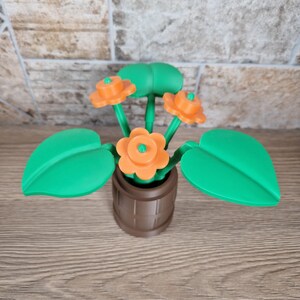 May include: A 3D-printed decorative flower arrangement featuring orange flowers with green centers and stems, and large green leaves. The flowers are in a brown pot with a barrel design. The arrangement is set against a brick wall.