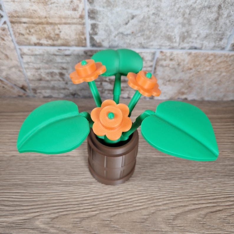 May include: A 3D-printed decorative flower arrangement featuring orange flowers with green centers and stems, and large green leaves. The flowers are in a brown pot with a barrel design. The arrangement is set against a brick wall.