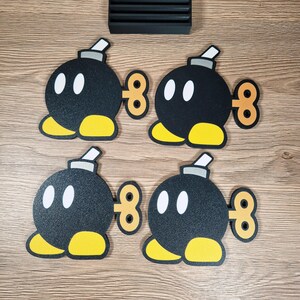 May include: Four black Bob-omb character coasters with white eyes, yellow feet, and a wind-up key design. A black rectangular coaster holder is also present. The coasters are designed for use with beverages.