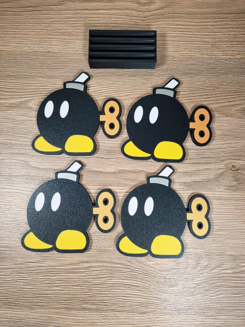 May include: Four black Bob-omb character coasters with white eyes, yellow feet, and a wind-up key design. A black rectangular coaster holder is also present. The coasters are designed for use with beverages.