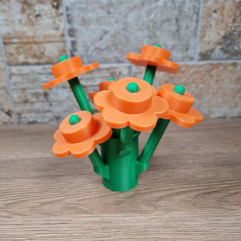 May include: A bouquet of orange flower-shaped objects with green stems and centers. The flowers are arranged in a green vase-like base. The background is a light brown wooden surface and a textured stone wall.