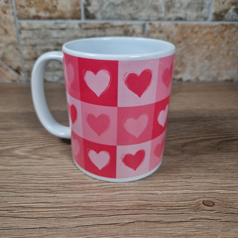 May include: White ceramic mug with a handle, featuring a repeating pattern of pink and red hearts in a checkerboard design. The mug is cylindrical and sits on a wooden surface.