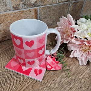 May include: A white ceramic mug with a pink heart pattern, matching coaster, and heart-shaped keychain. The mug and coaster feature a checkerboard design with various pink hearts. A bouquet of pink and white flowers is in the background.