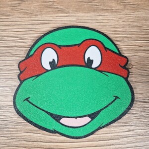 Puede incluir: Primer plano de la cara de una Tortuga Ninja, verde, roja y blanca. El personaje tiene una cabeza verde, una m&aacute;scara roja, ojos blancos y una boca sonriente.