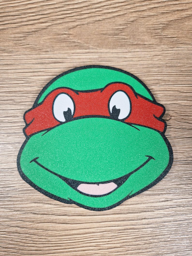 Puede incluir: Primer plano de la cara de una Tortuga Ninja, verde, roja y blanca. El personaje tiene una cabeza verde, una m&aacute;scara roja, ojos blancos y una boca sonriente.