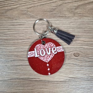 May include: A round red keychain with a white "Love" design and a gray tassel. The word "Love" is in white block letters over a pink leopard print heart. The keychain has a silver ring.