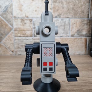 May include: A gray and black robot toy with a circular head and a rectangular body. The body features a red and white design. The robot has articulated arms and a circular base. The toy is made of plastic and sits on a wooden surface.