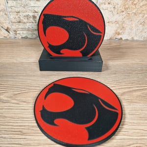 May include: Two round coasters with a red background and a black panther head design. One coaster is in a black holder, and the other is lying flat. The coasters are on a wooden surface.