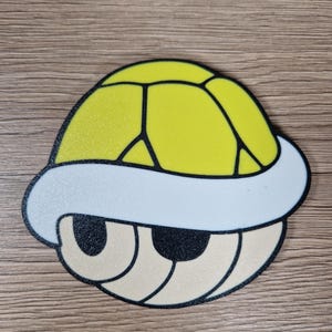K&ouml;nnte beinhalten: Eine gelbe Koopa Troopa-Schale mit wei&szlig;em Rand und schwarzen Umrissen. Die Schale hat eine strukturierte Oberfl&auml;che und ein cartoonartiges Design. Die Augen sind beige mit schwarzen Umrissen. Das Design befindet sich auf einer Holzoberfl&auml;che.