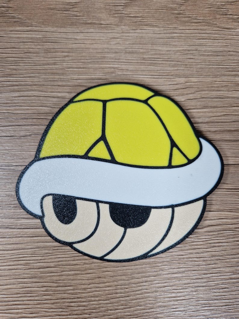 K&ouml;nnte beinhalten: Eine gelbe Koopa Troopa-Schale mit wei&szlig;em Rand und schwarzen Umrissen. Die Schale hat eine strukturierte Oberfl&auml;che und ein cartoonartiges Design. Die Augen sind beige mit schwarzen Umrissen. Das Design befindet sich auf einer Holzoberfl&auml;che.