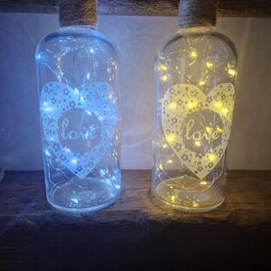 May include: Two glass bottles with a heart design and the word "love" on them. The bottles are filled with white fairy lights and have a rustic twine wrapped around the neck.