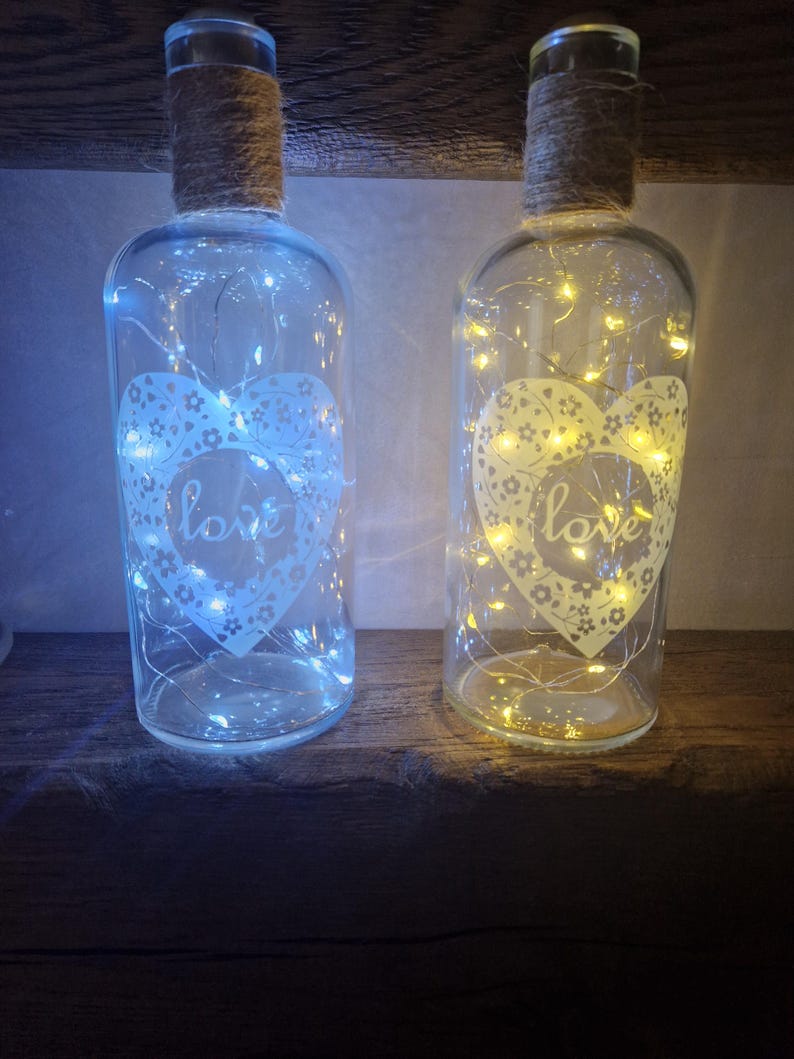 May include: Two glass bottles with a heart design and the word "love" on them. The bottles are filled with white fairy lights and have a rustic twine wrapped around the neck.