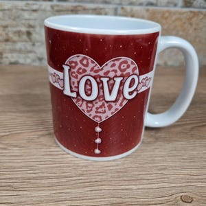 May include: A white ceramic mug with a red background and a heart-shaped design. The heart features a leopard print pattern and the word "Love" in white. The mug has a white handle and a white interior.