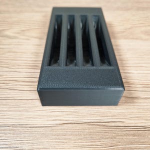 K&ouml;nnte beinhalten: Ein rechteckiges, dunkelgraues 3D-gedrucktes Objekt mit vier vertikalen Schlitzen. Das Objekt hat eine strukturierte Oberfl&auml;che und befindet sich vor einem hellbraunen Holzgrund. Das Design deutet auf ein Aufbewahrungs- oder Organisationsprodukt hin.
