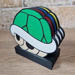 K&ouml;nnte beinhalten: Ein Satz bunter Schildkr&ouml;tenpanzer-Untersetzer. Die Untersetzer sind gr&uuml;n, rot, blau und gelb, mit einem wei&szlig;en und schwarzen Rand. Sie sind auf einem schwarzen rechteckigen Sockel gestapelt. Das Design erinnert an eine beliebte Videospiel-Figur.