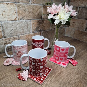 May include: Four white ceramic mugs with heart and "Love" designs, each with a matching coaster and heart-shaped keychain. The mugs are red and pink, with various patterns including hearts and the word "Love". A vase of pink and white flowers is in the background.