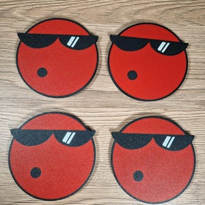 May include: Four red circular coasters with black sunglasses and a small black dot. The coasters are arranged in a square formation and are on a wooden surface. The sunglasses have white reflective lenses.