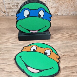Puede incluir: Juego de posavasos de las Tortugas Ninja. Cada posavasos presenta una cara de tortuga verde con una m&aacute;scara de color: azul, naranja, verde y rojo. Los posavasos son redondos y se colocan en un soporte negro. El fondo es una superficie de madera.