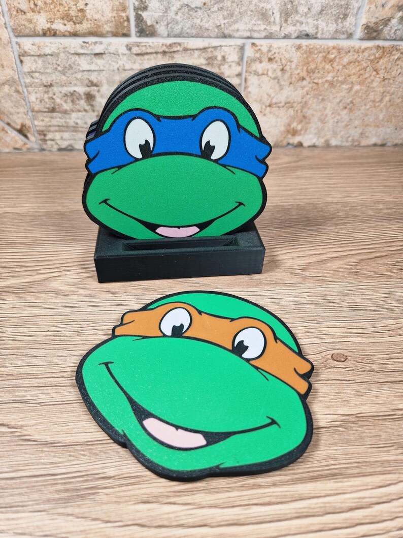 Puede incluir: Juego de posavasos de las Tortugas Ninja. Cada posavasos presenta una cara de tortuga verde con una m&aacute;scara de color: azul, naranja, verde y rojo. Los posavasos son redondos y se colocan en un soporte negro. El fondo es una superficie de madera.