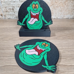 May include: Black coaster featuring a green cartoon ghost with a pink tongue and a wide grin. The coaster is round and has a black base. A second coaster is displayed in a black stand. The design is reminiscent of the Ghostbusters franchise.