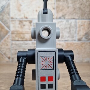 May include: A gray and black robot toy with a cylindrical head and a rectangular body. The body features a control panel with red and black squares and a sunburst design. The arms are black and articulated, and the toy stands on a black base.