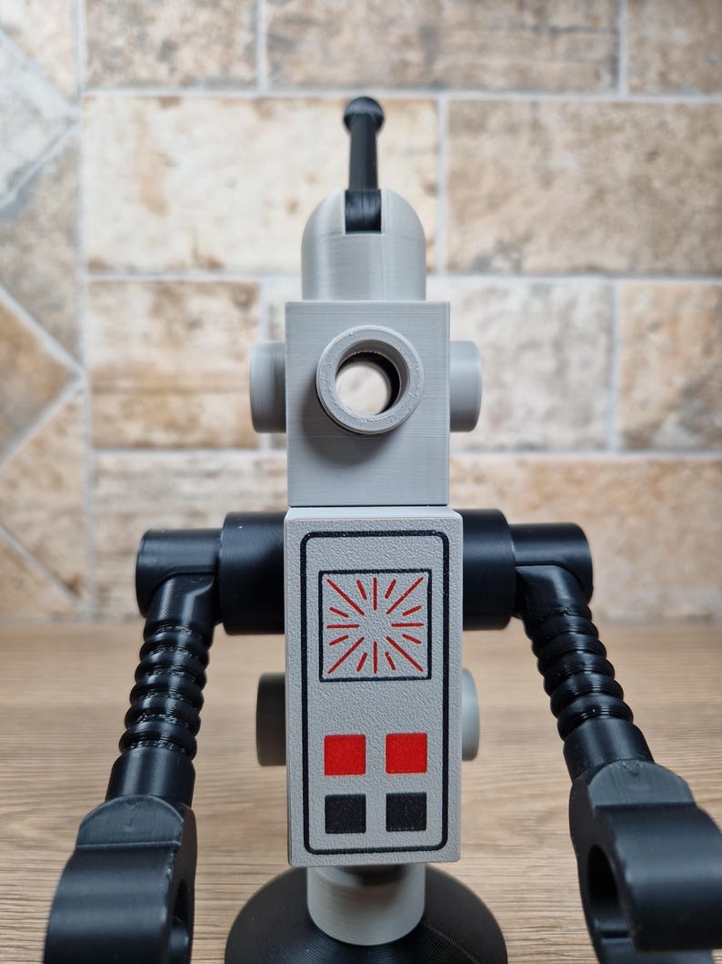 May include: A gray and black robot toy with a cylindrical head and a rectangular body. The body features a control panel with red and black squares and a sunburst design. The arms are black and articulated, and the toy stands on a black base.