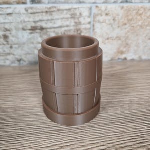 May include: A brown, barrel-shaped container with vertical lines and horizontal bands. The container has a cylindrical opening at the top and a flat base. The object is made of a solid material and sits on a wooden surface.