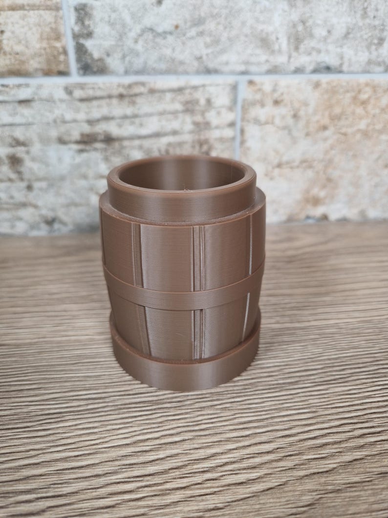 May include: A brown, barrel-shaped container with vertical lines and horizontal bands. The container has a cylindrical opening at the top and a flat base. The object is made of a solid material and sits on a wooden surface.