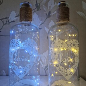 May include: Two clear glass bottles with a white heart design and the word "love" inside. The bottles are decorated with string lights and a brown twine around the top.