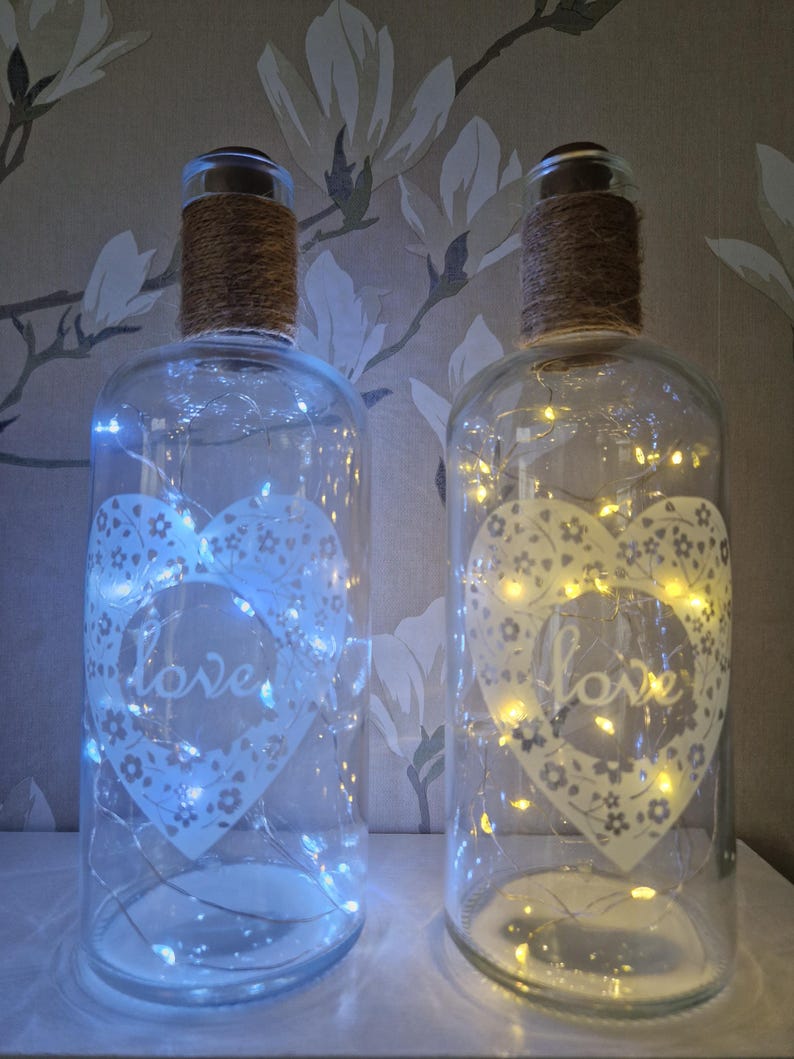 May include: Two clear glass bottles with a white heart design and the word "love" inside. The bottles are decorated with string lights and a brown twine around the top.
