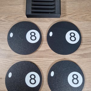 May include: A set of four black coasters with a white number 8 and a white dot, resembling a billiard ball. A small black rectangular holder with horizontal slots is also present. The items are arranged on a light brown wooden surface.