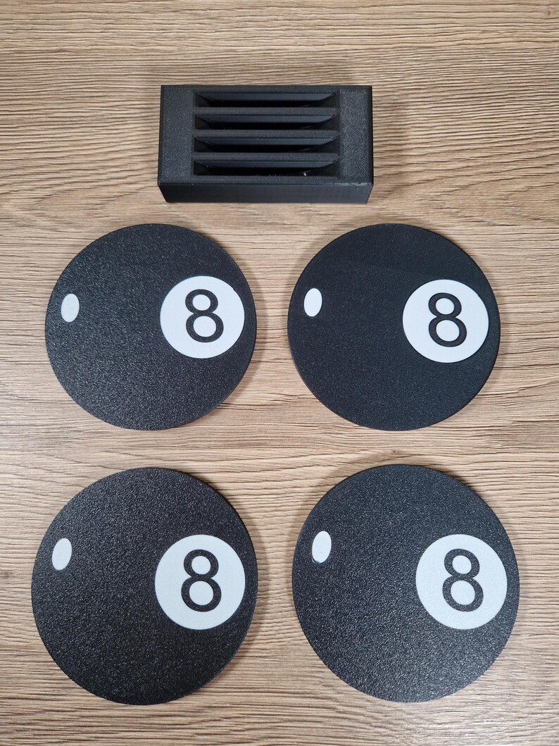 May include: A set of four black coasters with a white number 8 and a white dot, resembling a billiard ball. A small black rectangular holder with horizontal slots is also present. The items are arranged on a light brown wooden surface.