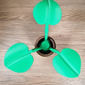 May include: A 3D-printed decorative plant with three bright green leaves. The leaves are heart-shaped and attached to a brown pot. The plant is made of plastic and sits on a wooden surface.