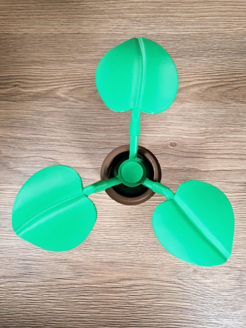 May include: A 3D-printed decorative plant with three bright green leaves. The leaves are heart-shaped and attached to a brown pot. The plant is made of plastic and sits on a wooden surface.